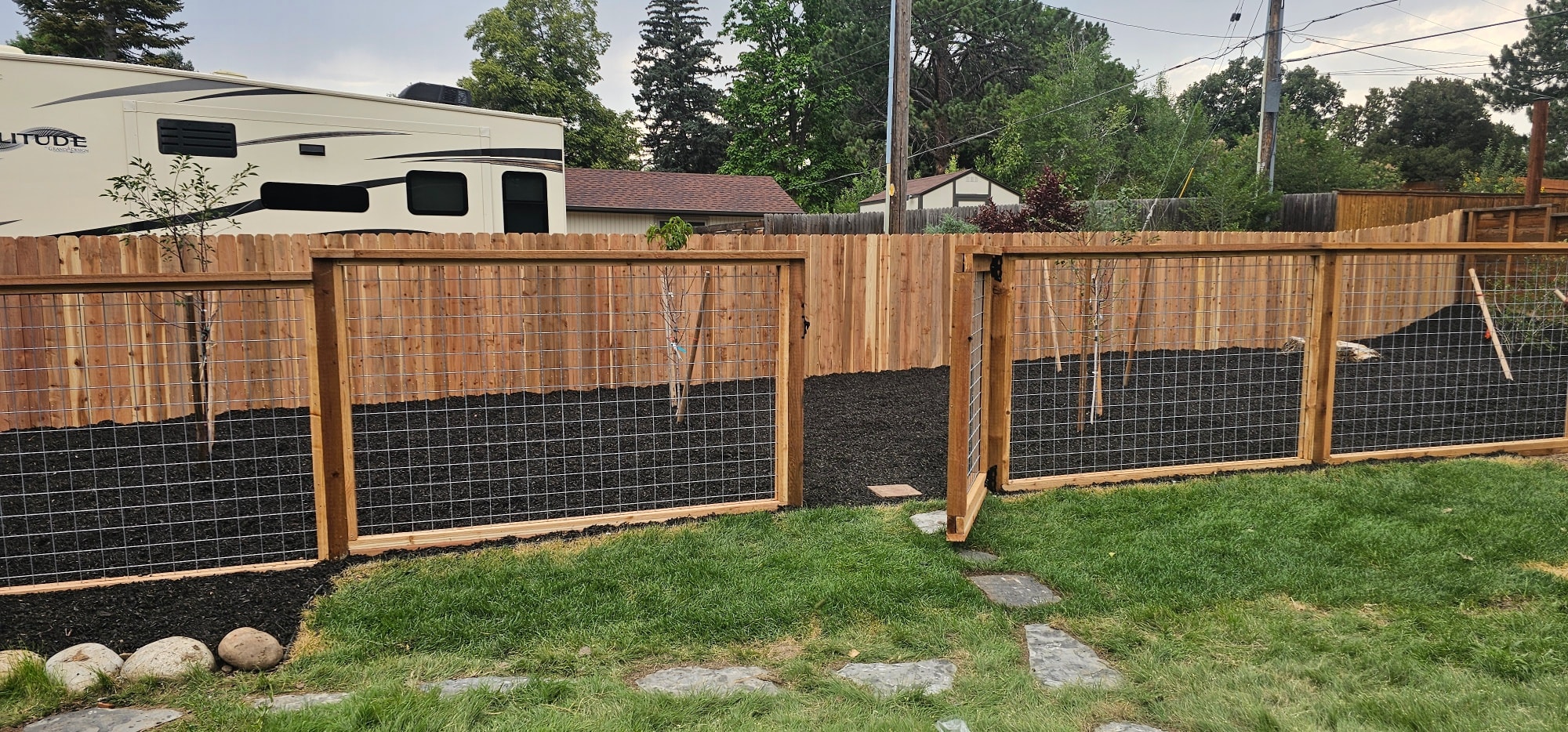 Wire mesh garden fence with a gate separating a grass lawn from a mulched planting area with young trees.