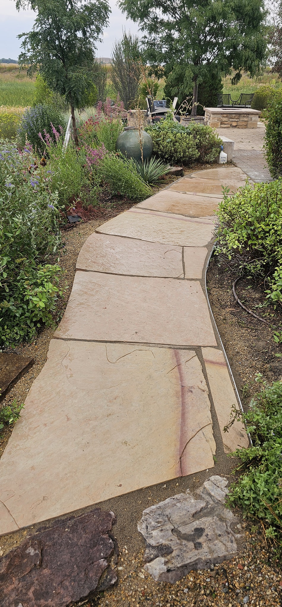 Flagstone garden path winding through lush landscaping with flowers and a stone fire pit area.