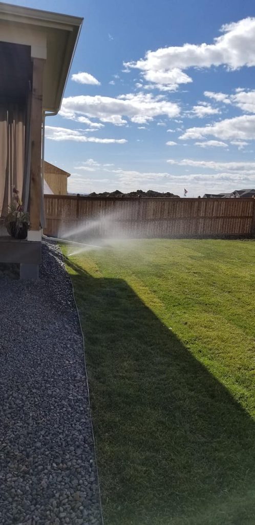 Backyard sprinkler system watering a green lawn under a bright blue sky with scattered clouds.