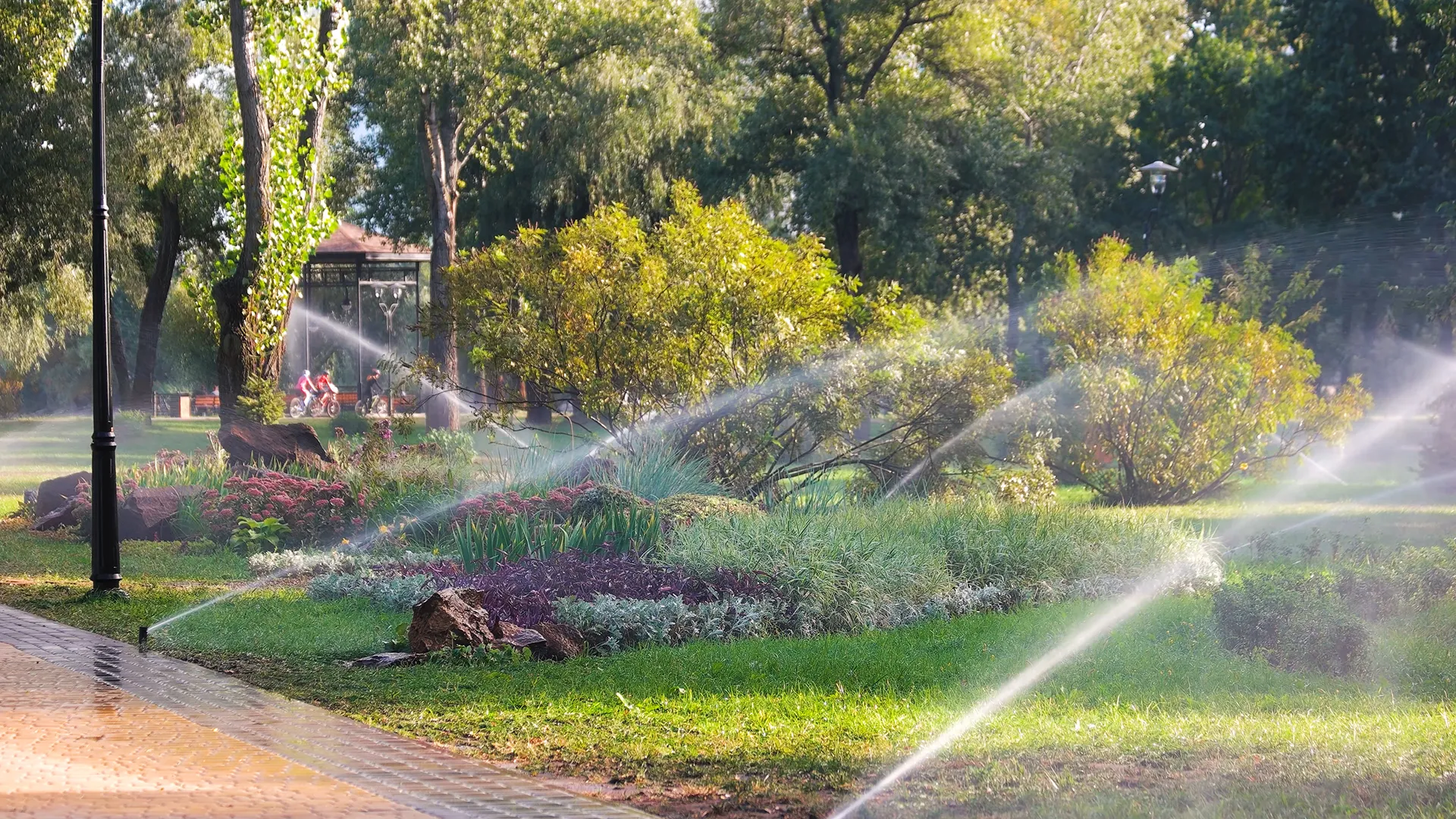 Automated sprinkler system misting a lush public park with flower beds and large trees in the morning light.